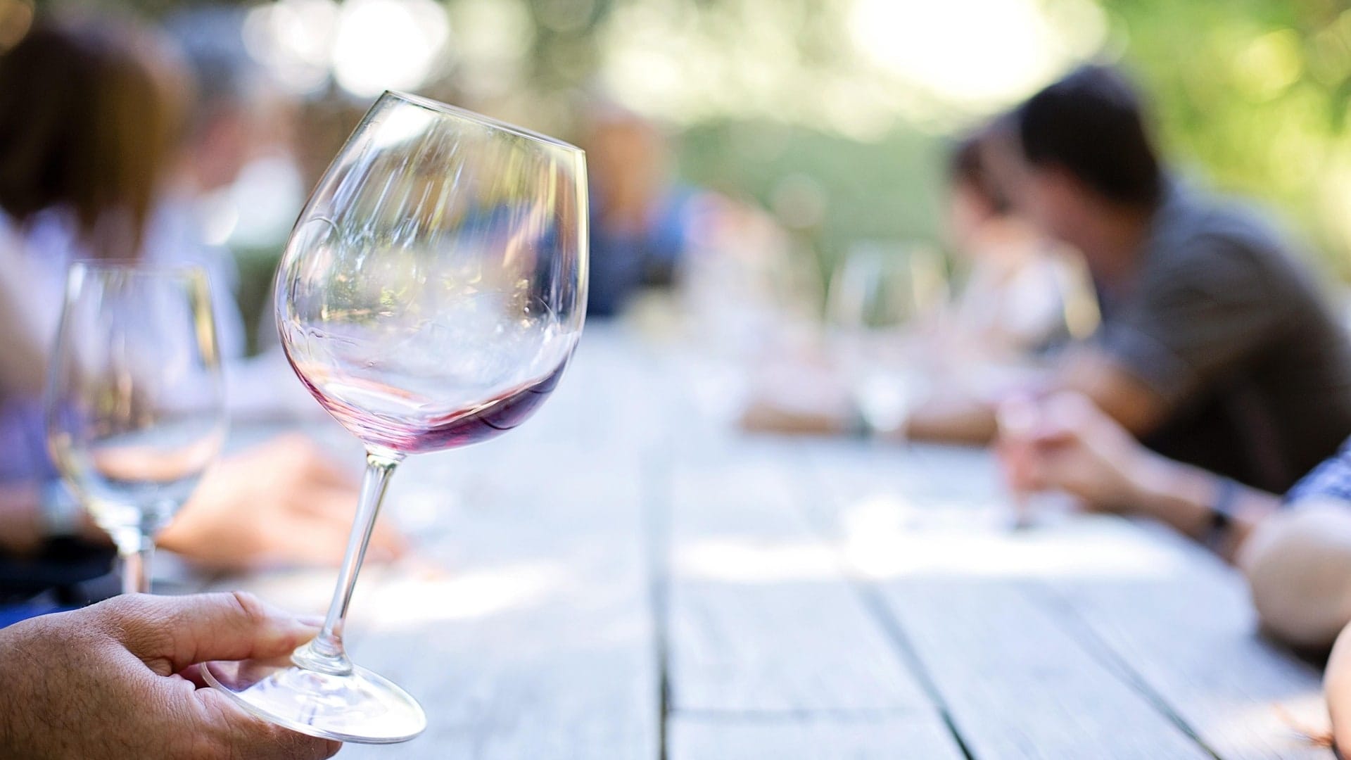 A glass of red held at a long outdoor table with people talking beyond it
