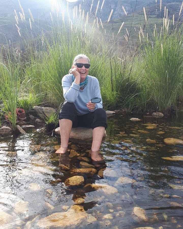 Angie Bradley laughing with her feet in a mountain stream, fynbos and cliffs behind her